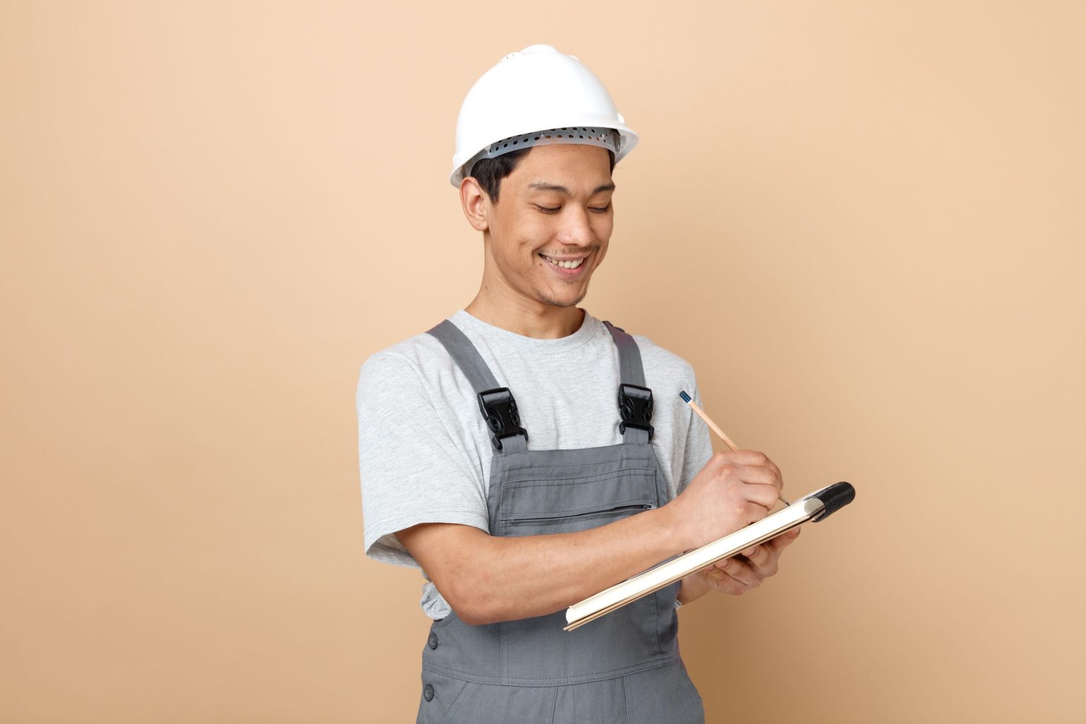 smiling-young-construction-worker-wearing-safety-helmet-uniform-writing-with-pencil-notepad